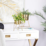 Styled interior scene showing two gold metal planters with greenery on a white desk under framed artwork