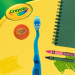 Blue Crayola toothbrush and orange holographic cap styled on yellow background with crayons and notebook props