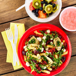 White plastic fork, knife and spoon beside a red bowl of pasta and fruit bowl on a wooden table