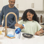 Lifestyle photo of a child using a Dawn Powerwash spray to clean a frying pan at a kitchen sink while an adult watches.