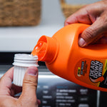 Close-up of hands pouring orange Dead Down Wind laundry detergent from bottle into a white measuring cap above a washer