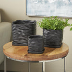 Three black wavy-ribbed ceramic planters displayed on a wooden side table with plant in one pot