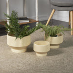 Three cream ribbed ceramic planters styled on a living room floor with two containing fern-like plants and a chair nearby.