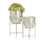 Two brushed silver metal planters on stands, one planted with green flowers, on white background