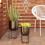 Two DecMode metal planters on stands placed on a patio with succulents in the pots and a wooden slat wall behind