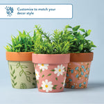 Three decorated terracotta pots with painted floral and leaf designs, filled with green foliage on a light blue background.