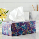 Close-up of a decorative rectangular tissue box on a countertop with a white tissue pulled out of the top and blurred background elements