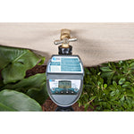 Digital water timer mounted on an outdoor brass spigot above garden plants, showing LCD and control buttons