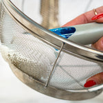 Close-up of a Dish Daddy brush with blue handle cleaning a metal mesh strainer, red-painted fingernails visible.