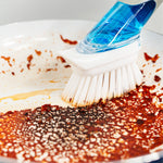 Dish Daddy brush with blue-handle attachment scrubbing burnt-on food from a frying pan in a close-up action shot.
