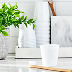 White disposable bathroom cup in foreground with stacked cups, a toothbrush, and a small plant in a bathroom styling scene.