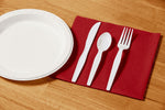 Place setting with white paper plate and disposable white knife, spoon and fork on a red napkin on a wood surface