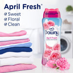 Downy April Fresh bottle shown beside a folded stack of pastel laundry and pink flowers in a laundry room scene