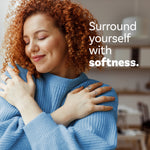 Lifestyle photo of a smiling woman hugging a soft blue sweater with text 'Surround yourself with softness.'
