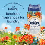 Downy Citrea scent booster bottle set against a sky-and-meadow background with floral accents and promotional boutique fragrance text.