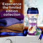 Styled lifestyle image of Downy Cuddle Weather bottle beside folded purple and brown sweaters and window backdrop with 'limited edition' text.