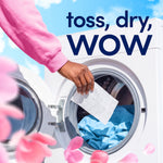 Hand placing a dryer sheet into a front-loading dryer drum with blue laundry inside and pink petals in the foreground.