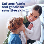 Woman and child smiling by a laundry machine, child holding a white towel, with text 'Softens fabric and gentle on sensitive skin'.