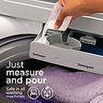 Hand pouring Downy fabric softener from the cap into a washing machine dispenser over a folded purple towel.