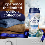 Lifestyle scene with folded sweaters and a Downy Cozy Collection Serene Snowfall scent booster bottle by a washer.