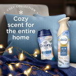 Cozy home scene showing Downy Cozy Collection Serene Snowfall bottle beside a Febreze Luxe spray on a pillow and lights.