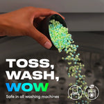 Hand pouring green and blue scent beads from a measuring cup into a washing machine with 'Toss, Wash, Wow' text.