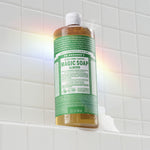 Dr. Bronner's almond liquid soap bottle on a tiled shower ledge with a soft rainbow light reflection.