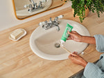 Person dispensing Dr. Bronner's spearmint toothpaste onto a toothbrush over a bathroom sink with soap dish and plant nearby