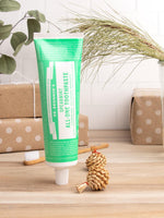 Lifestyle scene with a standing Dr. Bronner's spearmint toothpaste tube on a wooden surface beside a toothbrush and wrapped gifts