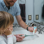 Smiling adult holding a tube of Dr. Brown's baby toothpaste while preparing to apply it to a child's toothbrush at the sink