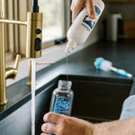 Close-up of Dr. Brown's Bottle & Dish soap being poured into a clear baby bottle over a kitchen sink, bottle brush visible in the background