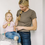 Child sitting on counter brushing with a yellow toddler toothbrush while an adult watches nearby