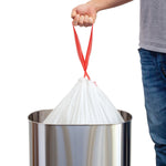 Hand lifting a tied white drawstring trash bag by red drawstrings above a stainless steel trash can