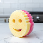 Close-up of a smile-faced dual-layer scrub sponge with yellow foam front and pink scrub back on a kitchen countertop
