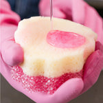 Close-up of a dual-sided scrub sponge with pink foam being applied and liquid poured onto it