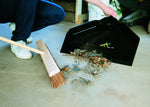 Close view of a black dust pan on concrete collecting dirt and leaves while a broom is positioned nearby