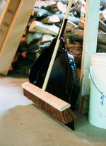 Black utility dust pan leaning against stacked firewood with a broom resting across the pan on a concrete floor