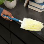 Hand holding a blue-handled duster with pale yellow refill cleaning a black countertop near books and a plant