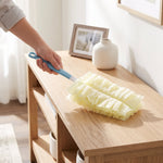 Hand using a blue-handled duster with a fluffy pale yellow refill to dust a wooden console table in a bright room