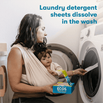 Woman carrying a baby in a sling loads an ECOS detergent sheets box into a front-load washing machine with product text above