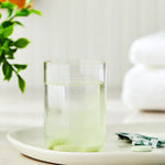 Glass of water with a green effervescing denture cleanser tablet dissolving on a white tray in a bathroom-like setting