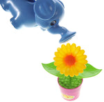 Blue elephant-shaped watering can positioned above a single pink pot with a yellow plastic sunflower on white background.