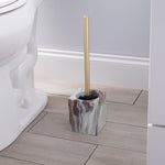 Gold-handled toilet brush standing in agate-patterned square holder placed on a bathroom floor beside a toilet.