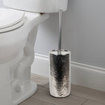 Chrome hammered ceramic toilet brush holder placed on bathroom floor next to toilet, chrome handle visible