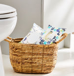 Lifestyle shot of two Equate Flushable Wipes packs in a woven basket placed on a bathroom floor next to a toilet.