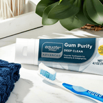 Toothpaste tube labeled Equate Gum Purify Deep Clean on a counter with a toothbrush and green plant in background