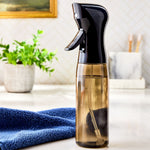 Amber continuous spray bottle with black top shown on a bathroom countertop beside a blue towel and plant