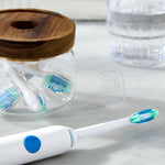 Close-up lifestyle shot of an Equate electric toothbrush head attached to a handle with extra heads in a glass jar on a countertop