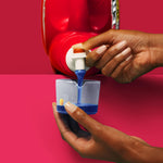 Hands dispensing blue ERA liquid detergent from a spout into a clear measuring cup on a red surface