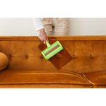 Hand holding green Evercare pet hair remover brushing a brown upholstered sofa, lifestyle usage shot